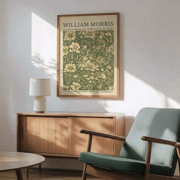 May include: Framed William Morris botanical poster with green floral pattern hanging above wooden credenza. Mid-century modern living room setting with sage green upholstered armchair, white ceramic table lamp, and round wood coffee table. Natural sunlight creates geometric shadows on white wall. Vintage-inspired exhibition art print in thin brown frame showcases intricate leaf and flower motifs in muted green tones.