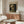 May include: Van Gogh museum poster of skull with cigarette artwork displayed above walnut wood credenza in minimalist living room with white curved chair, glass coffee table, and modern lamp. Poster shows skeleton head with burning cigarette against dark background in vintage exhibition style with cream border.