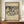 May include: A framed vintage-style poster featuring Wassily Kandinsky's abstract black ink artwork from 1911, displayed on a wooden credenza beside a small white ceramic vase and stacked books. The cream-colored print shows bold, flowing abstract forms and includes text referencing The National Gallery of Denmark. The poster has an aged, beige background with a thin brown frame, set against white wainscoted walls in a minimalist interior setting.