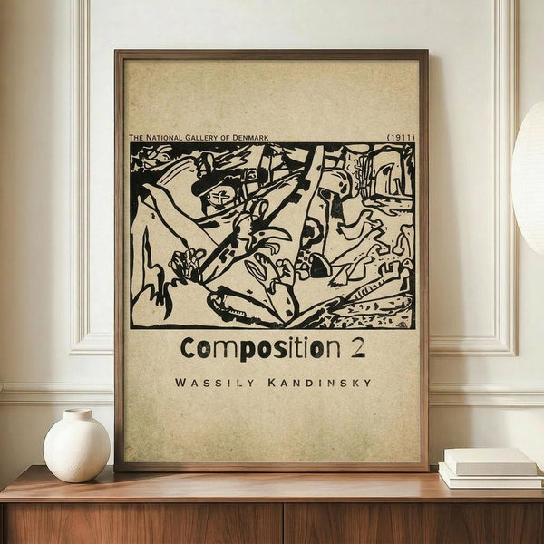 May include: A framed vintage-style poster featuring Wassily Kandinsky's abstract black ink artwork from 1911, displayed on a wooden credenza beside a small white ceramic vase and stacked books. The cream-colored print shows bold, flowing abstract forms and includes text referencing The National Gallery of Denmark. The poster has an aged, beige background with a thin brown frame, set against white wainscoted walls in a minimalist interior setting.