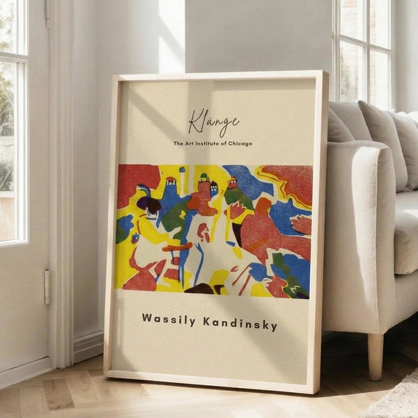 May include: A framed art poster leaning against a white wall next to a beige upholstered chair. The poster features a vibrant abstract artwork with bold colors including red, yellow, blue, green, and orange in flowing, organic shapes. The piece has handwritten-style text at the top and bottom on a cream background. Natural sunlight streams through nearby windows onto light wooden floors, creating a bright, modern living room setting.