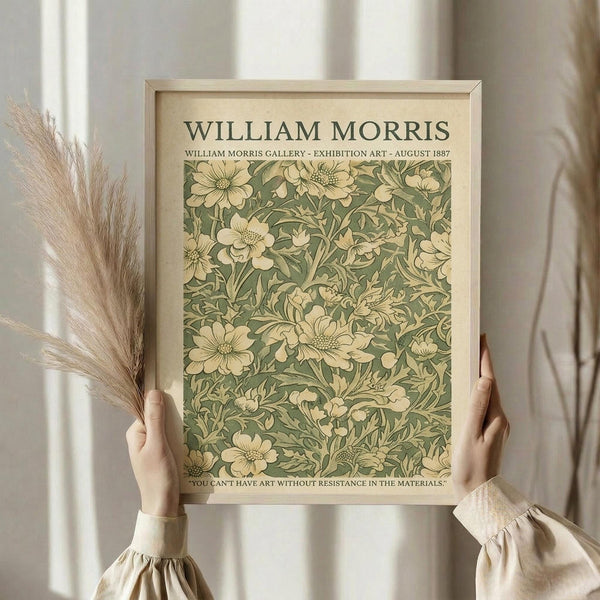 May include: Hands holding a framed vintage botanical art print featuring cream-colored flowers on sage green background with William Morris text at top. Light wooden frame, neutral beige curtains in background, dried pampas grass stems visible on left side. Soft natural lighting creates warm, minimalist aesthetic.