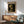May include: Van Gogh museum poster of skeleton smoking cigarette hanging on white wall above wooden credenza. Mid-century modern living room with sage green armchair, white table lamp, and natural sunlight casting shadows. Poster shows yellowed skull against dark background in vintage-style frame.