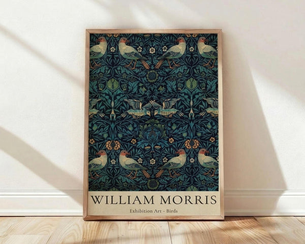 May include: Framed poster with William Morris-style pattern featuring birds and botanical elements in deep teal, green, and coral colors against dark navy background. Wood frame sits on light wooden floor against white wall with natural lighting creating soft shadows.