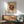 May include: Framed abstract art poster with burgundy geometric design featuring circular and linear elements on cream background, displayed above wooden sideboard with table lamp in modern living room setting with mint green upholstered chair and natural lighting casting shadows on white wall.