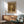 May include: Vintage-style poster featuring a brown bear in a sunlit forest with tall trees, titled "Wildwood Chronicles - 1974" and "Preserve Nature" in brown frame hanging above wood credenza with white lamp, next to mint green mid-century modern chair with wooden arms in bright room with geometric shadows on white wall.