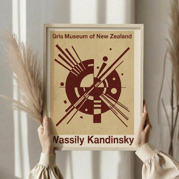 May include: Person in cream sweater holding framed abstract art poster featuring burgundy geometric circles, lines and dots on beige background, displayed in light wood frame against white curtained window, with dried wheat stems held in other hand