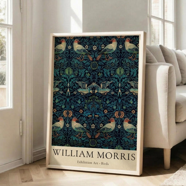 May include: Framed art print featuring William Morris-style pattern with colorful birds and botanical elements in deep blues, greens, and earth tones, displayed in light wooden frame leaning against white wall next to beige upholstered chair in bright, sunlit room with hardwood floors.