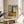 May include: Framed Van Gogh cypress tree poster displayed on round wooden side table next to beige upholstered chair. Print shows tall dark cypress against golden wheat field and swirling cloudy sky in post-impressionist style with green, yellow and blue tones. Thin wooden frame with cream matting. Soft natural lighting creates window shadows on white wall. Green plant leaves visible in corner.