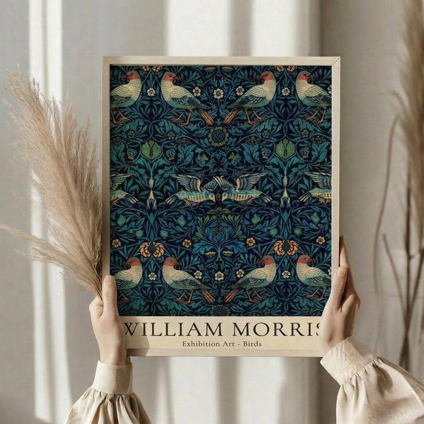 May include: Hands holding a framed art print featuring an intricate William Morris-style pattern with colorful birds and botanical motifs on a dark blue background. The print has a light wooden frame and shows repeating designs of parrots, owls in flight, leaves, and flowers in greens, blues, and earth tones. Dried pampas grass stems are visible to the left, and soft natural lighting creates shadows on neutral curtains in the background.