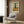 May include: Van Gogh-inspired poster featuring a tall dark cypress tree in a golden wheat field under swirling cloudy sky, displayed in light wood frame on warm brown wood-paneled wall above beige upholstered armchair with geometric patterned pillow, beside brass floor lamp and dark round side table with open book