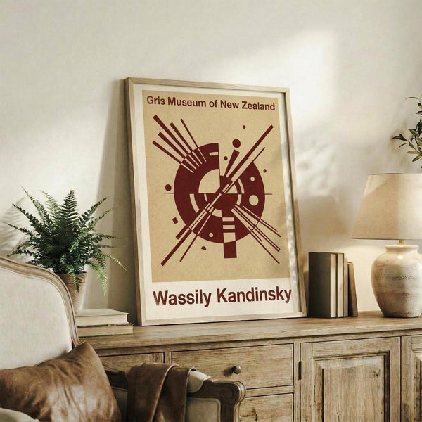 May include: Framed abstract art print with burgundy geometric design featuring radiating lines and circular elements on cream background, displayed on rustic wooden sideboard with books, ceramic vase, and fern plant in cozy living room setting with beige armchair and neutral walls