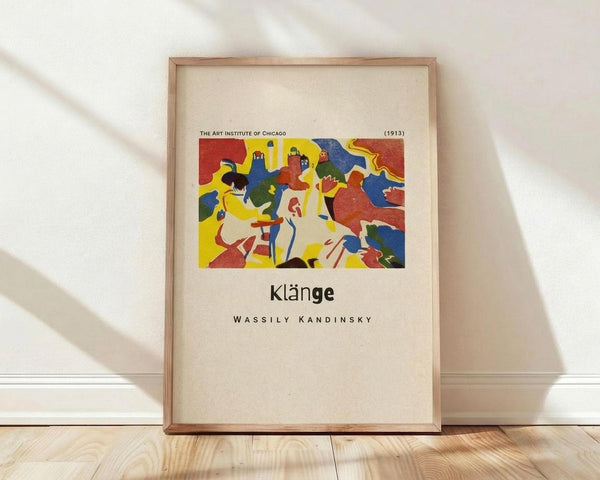 May include: Framed art print leaning against white wall on light wood floor. Print features colorful abstract artwork with red, yellow, blue, and green shapes depicting stylized figures. Text reads "The Art Institute of Chicago (1913)" at top, "Klänge" in center, and "Wassily Kandinsky" below. Natural wood frame with cream matting. Soft window light creates diagonal shadows across white fabric backdrop.