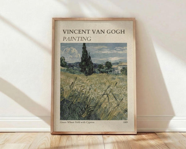 May include: Framed art print featuring Van Gogh's wheat field landscape with tall dark cypress tree, golden grain field in foreground, and cloudy sky. Print has cream-colored background with black text at top and bottom. Thin light wood frame leans against white wall on natural wood floor with soft window light creating shadows.