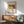 May include: Framed art print featuring Wassily Kandinsky's colorful abstract composition with bold red, blue, yellow, and green geometric shapes and figures. Print displays "Klänge" text and Art Institute of Chicago attribution. Shown in wood frame hanging above mid-century modern credenza with white lamp, alongside sage green upholstered chair with wooden arms. Warm sunlight creates dramatic shadows across white wall.