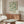 May include: Framed botanical poster with green leafy pattern on cream background displayed above wood sideboard in modern living room. Room features white curved chair, glass oval coffee table, brass floor lamp, and neutral walls with natural lighting.