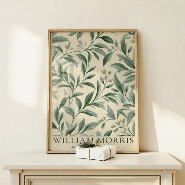 May include: A framed botanical art print featuring flowing green leaves and stems in William Morris style on cream background, displayed on a light wood console table with a small wrapped gift box beside it against a neutral wall.