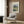 May include: Van Gogh poster print in light wood frame hanging on wood-paneled wall above beige upholstered chair with geometric pillow. Print shows wheat field landscape with cypress trees and mountains in blues, greens, and golden yellows. Modern reading corner with brass floor lamp and side table.