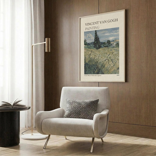 May include: Van Gogh poster print in light wood frame hanging on wood-paneled wall above beige upholstered chair with geometric pillow. Print shows wheat field landscape with cypress trees and mountains in blues, greens, and golden yellows. Modern reading corner with brass floor lamp and side table.