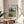 May include: Framed Van Gogh wheat field reproduction poster displayed on round wooden side table next to beige upholstered chair. Print shows golden wheat field with tall dark cypress tree and cloudy sky in post-impressionist style. Natural lighting creates soft shadows across neutral-toned room setting with plant leaves visible in corner.