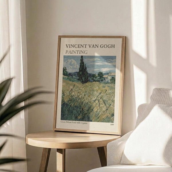 May include: Framed Van Gogh wheat field reproduction poster displayed on round wooden side table next to beige upholstered chair. Print shows golden wheat field with tall dark cypress tree and cloudy sky in post-impressionist style. Natural lighting creates soft shadows across neutral-toned room setting with plant leaves visible in corner.