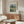 May include: Van Gogh reproduction poster featuring a golden wheat field with tall dark cypress tree and buildings under swirling sky, displayed in thin brown frame above mid-century walnut credenza with sliding doors. Modern living room setting with curved white chair, glass oval coffee table, and brass floor lamp creating warm natural lighting on white walls.