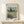 May include: Framed Van Gogh painting reproduction showing golden wheat field with tall dark cypress tree and rolling hills under cloudy sky, displayed on light wood console table beside small white gift box with natural twine and greenery, against cream colored wall