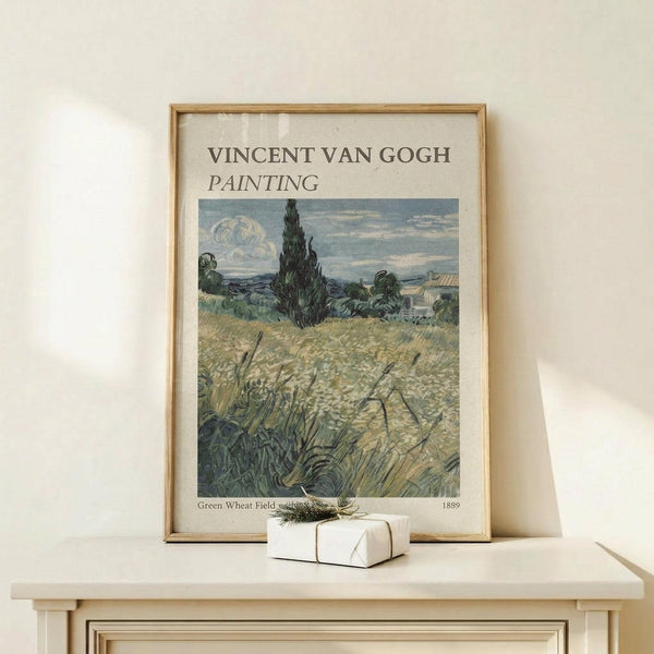 May include: Framed Van Gogh painting reproduction showing golden wheat field with tall dark cypress tree and rolling hills under cloudy sky, displayed on light wood console table beside small white gift box with natural twine and greenery, against cream colored wall
