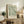 May include: Framed botanical art print with flowing green and cream leaf patterns displayed on rustic wooden sideboard alongside fern plant, stacked books, ceramic vase, and table lamp in neutral-toned living room setting