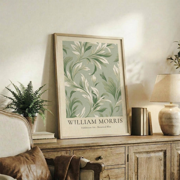 May include: Framed botanical art print with flowing green and cream leaf patterns displayed on rustic wooden sideboard alongside fern plant, stacked books, ceramic vase, and table lamp in neutral-toned living room setting