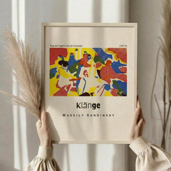 May include: Person in cream ruffled sleeve top holding framed art print against white curtained window, with dried wheat stems visible to left. Frame shows colorful abstract artwork with red, yellow, blue, and green flowing shapes and figures. Museum-style poster format with beige matting and thin wooden frame.
