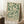 May include: Framed botanical poster with cream background featuring green leafy branches and buds in William Morris style, displayed on white fluffy rug against white wall corner. Coffee cup on wooden surface visible to left, beige blanket draped nearby. Natural window light creates soft shadows on wall.