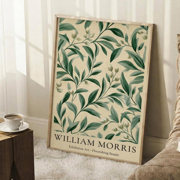 May include: Framed botanical poster with cream background featuring green leafy branches and buds in William Morris style, displayed on white fluffy rug against white wall corner. Coffee cup on wooden surface visible to left, beige blanket draped nearby. Natural window light creates soft shadows on wall.