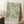 May include: Framed botanical poster with flowing green and cream leaf pattern on sage background, displayed in bright room corner next to wooden side table with white coffee cup and saucer, leaning against white paneled wall on textured cream rug with tan blanket visible at edge