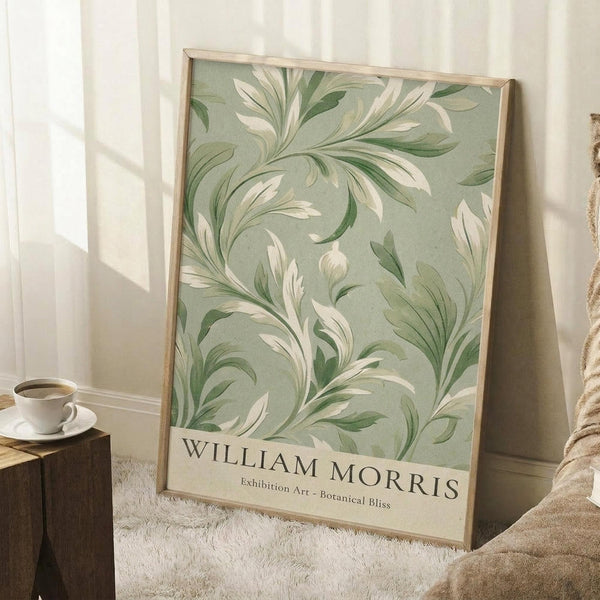 May include: Framed botanical poster with flowing green and cream leaf pattern on sage background, displayed in bright room corner next to wooden side table with white coffee cup and saucer, leaning against white paneled wall on textured cream rug with tan blanket visible at edge