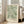 May include: Large framed botanical art print leaning against white wall next to beige upholstered chair. Features flowing green and cream acanthus leaf pattern on sage background with William Morris text below. Light wood frame, soft natural lighting from nearby windows, hardwood floors with area rug edge visible.