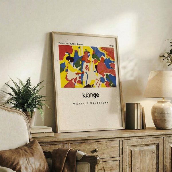 May include: Framed art print displayed on rustic wooden sideboard in living room setting. Abstract colorful artwork features bold red, blue, yellow shapes with geometric figures. Light wood frame with white matting. Scene includes cream upholstered chair, fern plant, stacked books, ceramic vase, and neutral wall background. Print shows vibrant modernist style with angular human forms and dynamic composition.