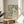 May include: Framed botanical art print with green leafy pattern on cream background, displayed on round wooden side table next to beige upholstered chair. Natural lighting creates soft shadows across neutral-toned room with white curtains and houseplant visible in corner.