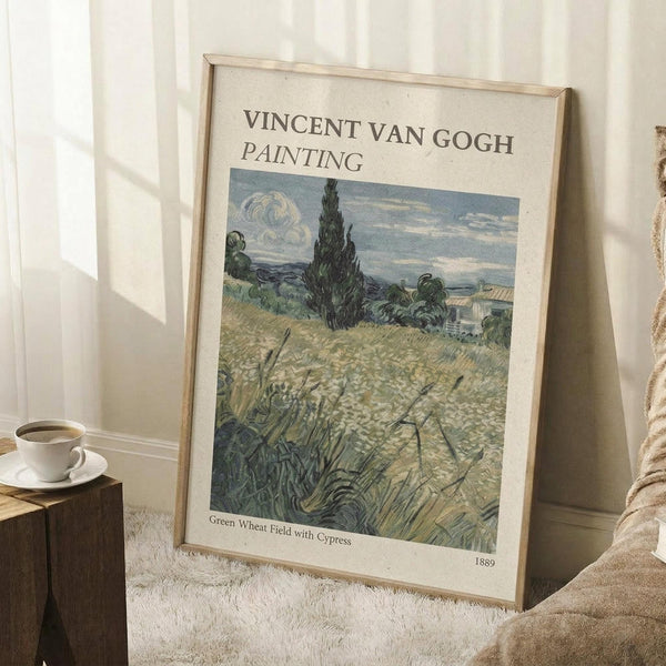 May include: Framed Van Gogh painting print leaning against white wall on textured cream rug. Artwork shows golden wheat field with tall dark cypress tree, rolling hills, and cloudy sky in post-impressionist style. Natural wood frame, displayed near wooden block with white coffee cup and beige blanket.