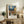 May include: Framed Van Gogh-inspired poster print displayed on rustic wood sideboard in cozy living room setting. Artwork shows Mediterranean village scene with swirling night sky, warm yellow buildings, and church spire. Natural wood frame with white matting. Scene includes fern plant, stacked books, ceramic vase, and cream upholstered furniture against neutral walls.
