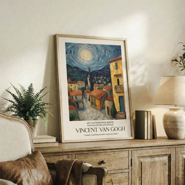 May include: Framed Van Gogh-inspired poster print displayed on rustic wood sideboard in cozy living room setting. Artwork shows Mediterranean village scene with swirling night sky, warm yellow buildings, and church spire. Natural wood frame with white matting. Scene includes fern plant, stacked books, ceramic vase, and cream upholstered furniture against neutral walls.