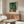 May include: A framed Van Gogh poster print of swirling cypress trees in vibrant greens and blues, displayed on a warm wood credenza in a minimalist living room with cream walls, white curved chair, glass coffee table, and modern brass floor lamp.