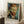 May include: Framed art print of Van Gogh's self-portrait featuring his distinctive red beard, blue eyes, and swirling brushstroke style in blues and greens, displayed leaning against white wall corner on white textured rug next to wooden side table with white coffee cup and saucer, natural window light creating soft shadows.