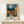 May include: Framed poster print displayed on light wood console table against white wall. Artwork shows Mediterranean village scene with swirling night sky in Van Gogh style, featuring warm yellow buildings with red tile roofs, church tower, and dramatic blue sky with circular moon. Small white gift box with greenery sits beside the natural wood frame.