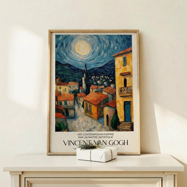 May include: Framed poster print displayed on light wood console table against white wall. Artwork shows Mediterranean village scene with swirling night sky in Van Gogh style, featuring warm yellow buildings with red tile roofs, church tower, and dramatic blue sky with circular moon. Small white gift box with greenery sits beside the natural wood frame.