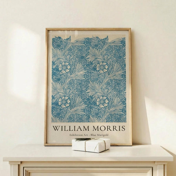 May include: A framed botanical poster featuring an intricate blue and cream floral pattern with marigold flowers and flowing foliage in William Morris's distinctive Arts and Crafts style. The print is displayed in a light wood frame on a cream-colored wooden console table against a neutral wall, with a small wrapped gift box placed in front.