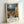 May include: Framed poster print leaning against white wall showing Van Gogh-style painting of Mediterranean village with swirling night sky, bright moon, terracotta rooftops, and yellow buildings. Features French text and artist attribution below artwork. Light wood frame with white matting, positioned on white surface with soft natural lighting creating gentle shadows.