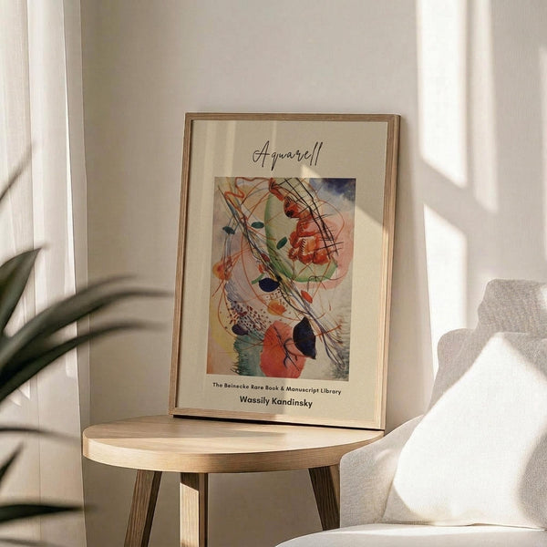 May include: Framed abstract watercolor print displayed on round wooden side table next to beige upholstered chair. Artwork features flowing organic shapes in orange, green, blue and black with curved lines and geometric elements. Natural wood frame with cream matting. Soft sunlight creates shadows across white wall. Green plant leaves visible in corner.