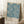 May include: Framed poster featuring an intricate blue floral pattern with marigold flowers and leafy foliage on cream background, leaning against white wall corner next to wooden side table with coffee cup, beige throw blanket visible at edge, natural window light creating soft shadows on white fluffy rug.