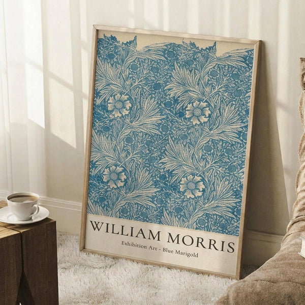 May include: Framed poster featuring an intricate blue floral pattern with marigold flowers and leafy foliage on cream background, leaning against white wall corner next to wooden side table with coffee cup, beige throw blanket visible at edge, natural window light creating soft shadows on white fluffy rug.